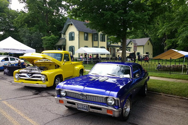 Two classic cars, one with an open hood, are parked in front of a yellow Victorian home with green trim.