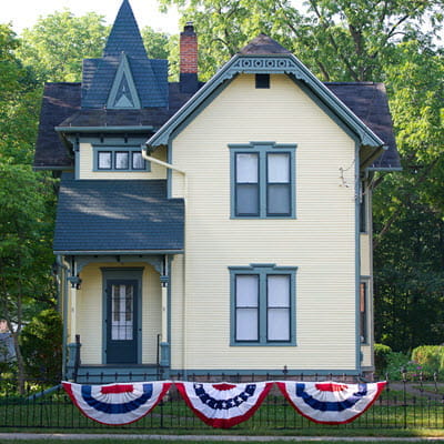 A 2-story Late Victorian home with yellow exterior with green trim sits behind a wrought-iron fence.