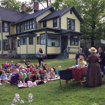 A group of children sit on a lawn outside a Victorian home; adults at a table in front of them hold historical artifacts.