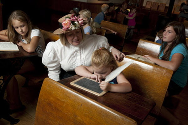 A women in historical clothing kneels next to a young girl; the girl is writing on a slate with a quill pen. 