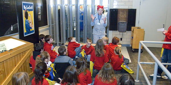 A woman in overalls and a red bandana talks to elementary students next to a World War II Jeep.