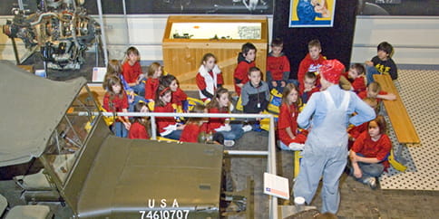 A woman in overalls and a red bandana talks to elementary students next to a World War II Jeep.