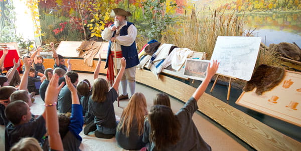 An elementary class listens to a man with a white beard and tri-corner hat talk in a museum exhibit. 