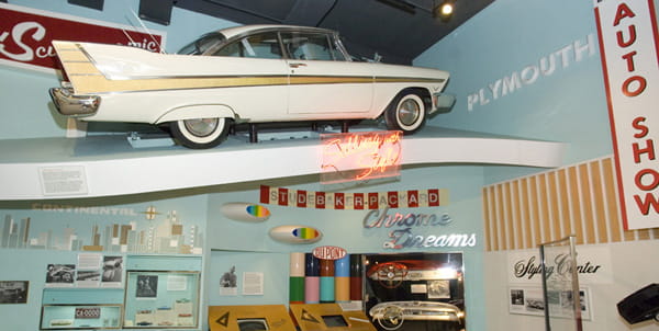 A white and gold car with large tailfins is suspended above an exhibit on 1950s car design. 
