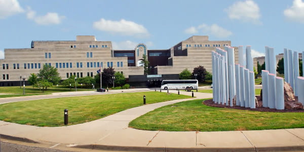 Exterior of 5-story limestone building with a white pine in the middle; a tour bus is parked in front a  large sculpture is in the foreground. 
