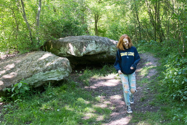 A teen girl walks along a dirt footpath. Large boulders are to her right. They are surrounded by forest.