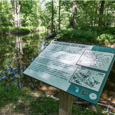 A sign about the Cass River on a trail. A muddy river flows in the background. 