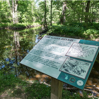 A sign about the Cass River on a trail. A muddy river flows in the background. 