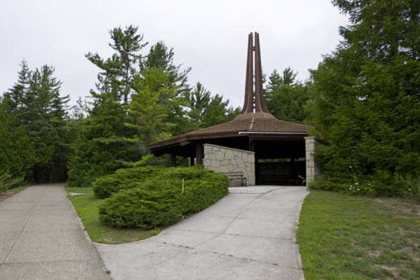 A stone 1-story pavilion with a brown shingled roof sits among conifer trees and shrubbery