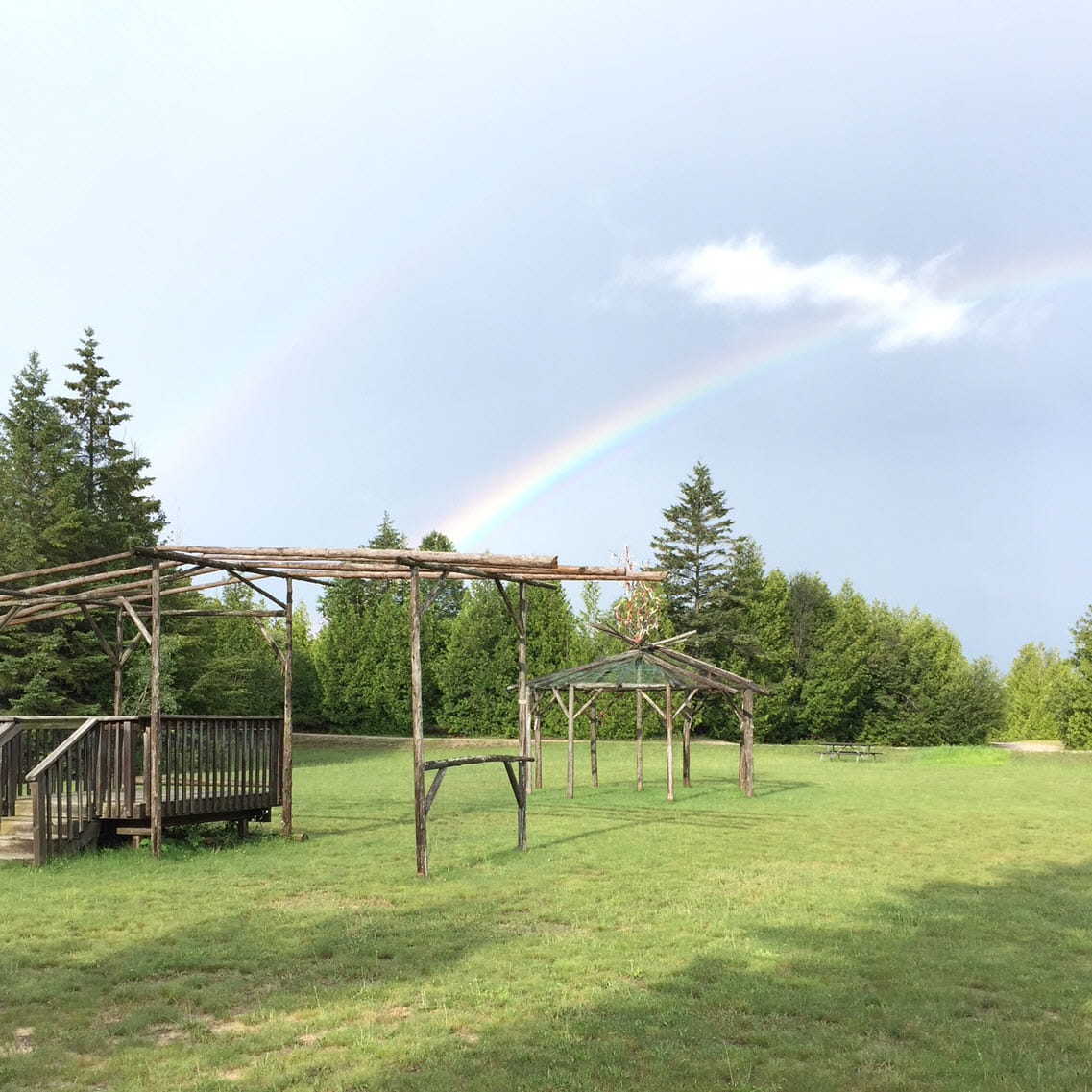 A double rainbow in the sky above an open field. Log structures dot the grounds. 