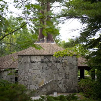 A stone 1-story pavilion with a brown shingled roof, viewed through conifer trees and shrubbery
