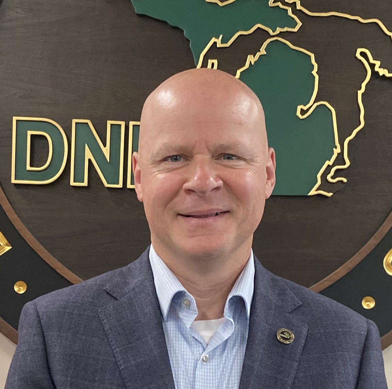headshot of man in a grey suit standing in front of a DNR sign