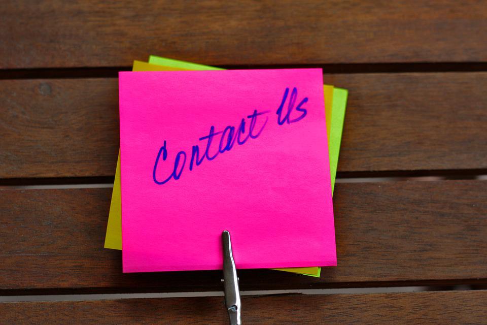 Small, square, pink paper note, held up with a clip in front of a brown wall, with handwriting that says contact us.