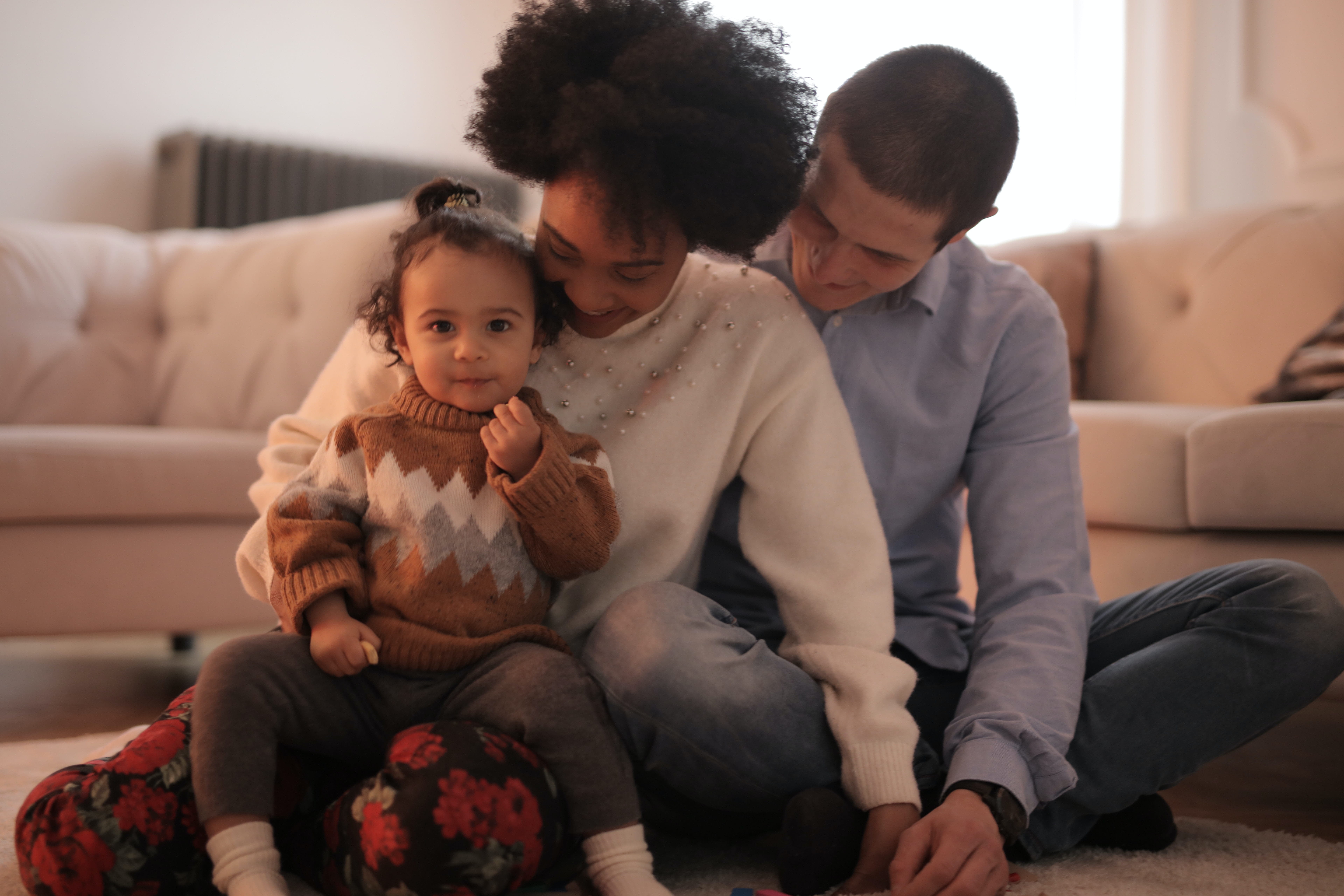 Two smiling adult caregivers and a toddler, sitting together on a living room floor, sharing a moment as a family.