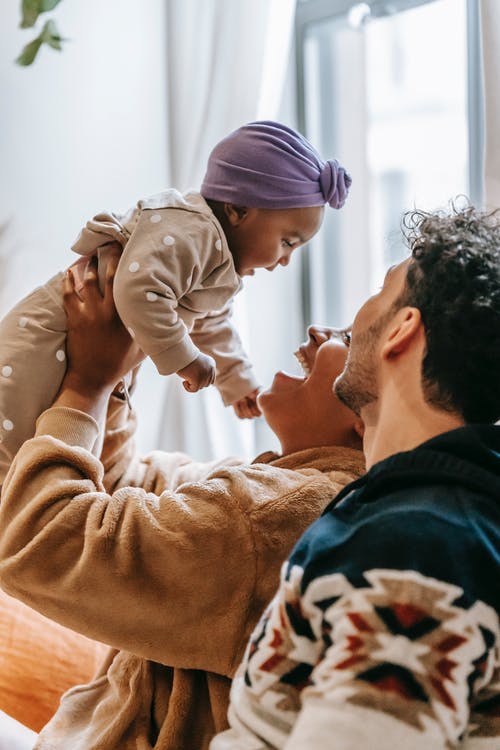 Two adults sitting next to each other, laughing and looking up at an infant being lifted by one of the adults.