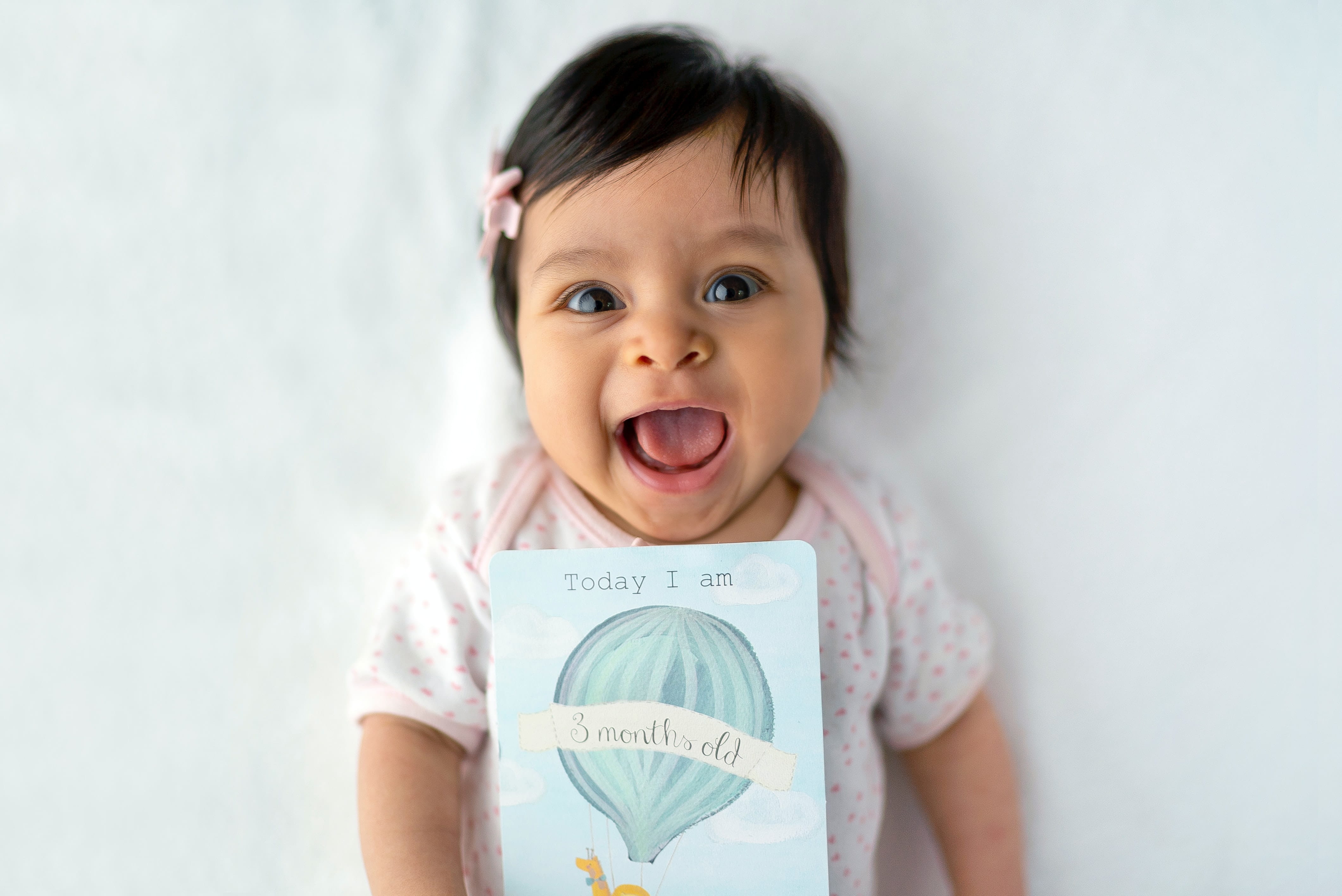 Infant with dark hair wearing a pink hairclip, laying on a white surface and looking at the camera, laughing.