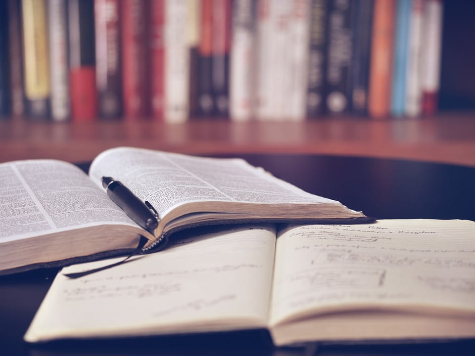 Two open books laying on a wooden desk with a row of books sitting upright in the background.