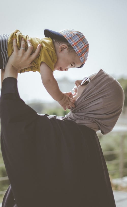 A person standing outdoors, lifting and smiling at a baby while looking into the baby's eyes.
