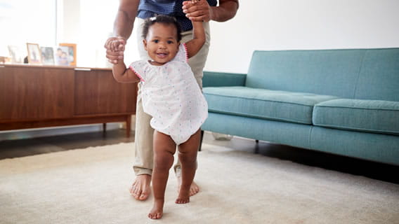parent holding infant hands to guide her as she takes steps