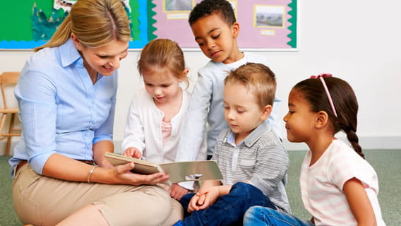 four preschoolers gathered around teacher while she reads and shows  them a book