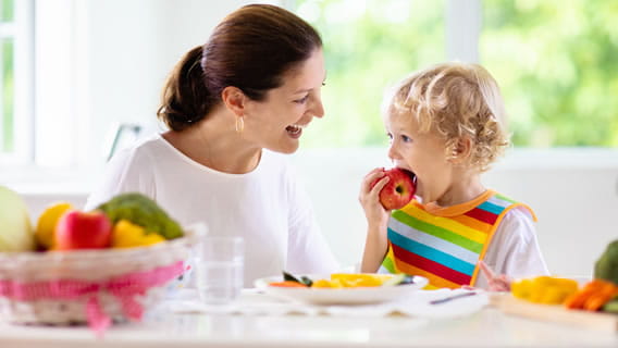 mom dining table smiling at preschool aged child taking a bite out of an apple