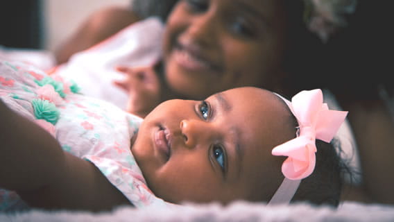 baby in dress and headband lying on bed while sister looks over her smiling