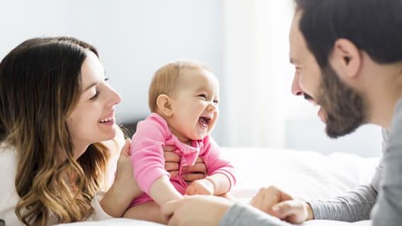 mom laying on bed smiling and holding infant up as dad smiles an touched baby's leg