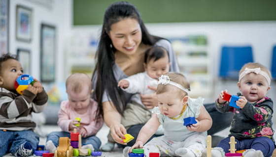provider holding infant while smiling and watching two other infants playing in care setting