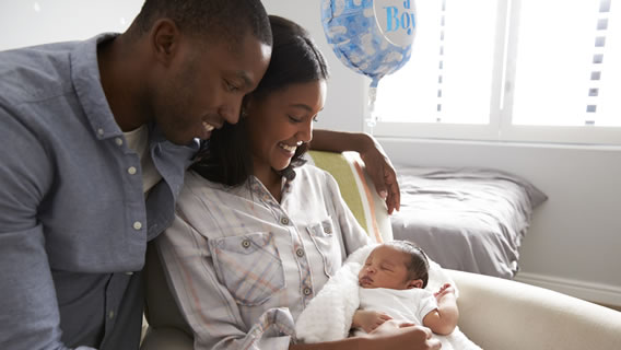mom holding baby with dad looking over her next to "it's a boy" balloon