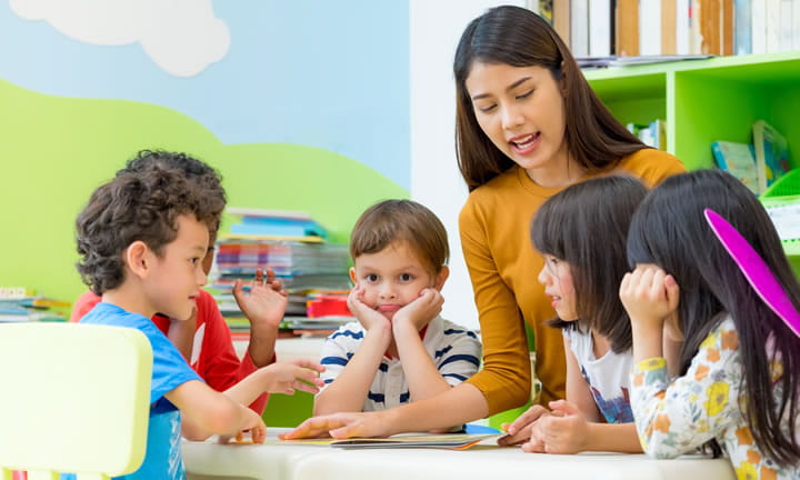 kindergarten teacher at table with five students reading a book to them