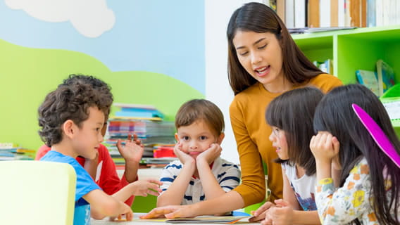 kindergarten teacher at table with five students reading a book to them