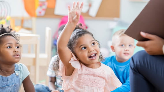 smiling preschool girl raising hand in classroom setting as teacher reads book to class