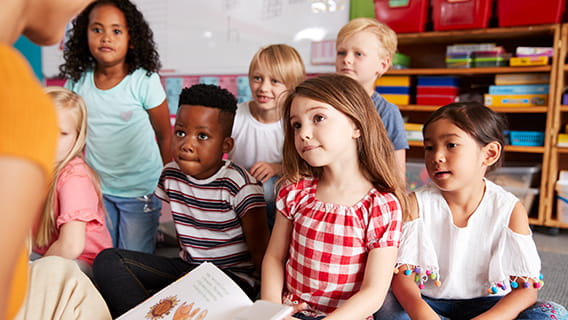 preschooler sitting on floor in classroom intently listening to teacher read a picture book