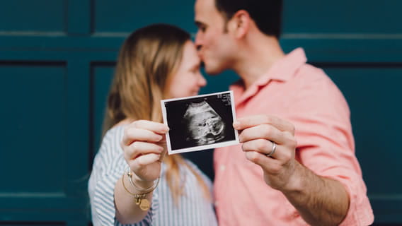 expectant parents holding out sonogram while dad kisses moms forehead