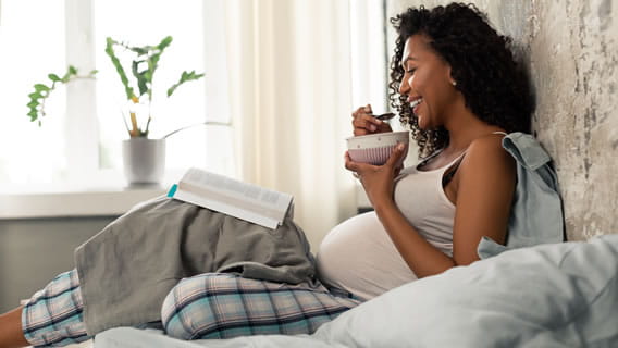 expectant mother eating from bowl sitting in bed while reading book