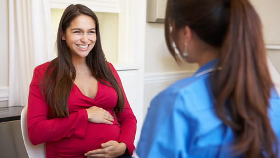 expectant mother holing belly and smiling while sitting and talking to nurse