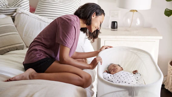 mom sitting on bed leaning over baby in bassinet