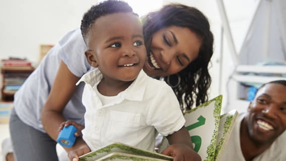 toddler holding book with mom smiling behind him supporting book