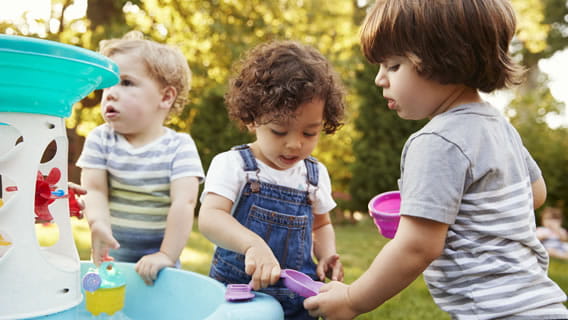 three toddlers outside playing with cups and shovels at a plastic toy fountain