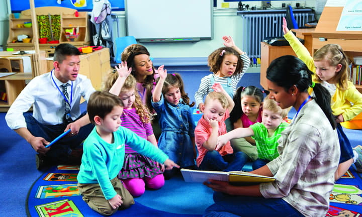 preschoolers sitting on floor in classroom raising their hands as provider reads to them