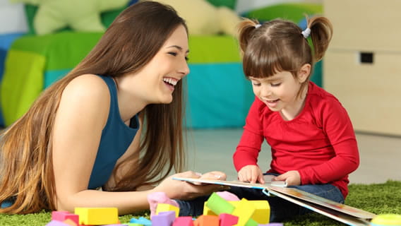 Preschooler in care setting sitting on the floor reading a book as smiling provider is propped on elbows beside her