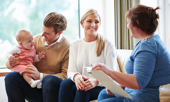 dad on couch holding and looking at baby and sitting next to mom as they listen to nurse