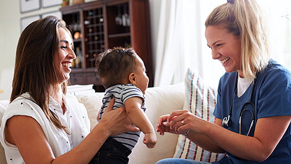 mom at home on couch holding baby out to nurse as nurse holds arms out to receive baby