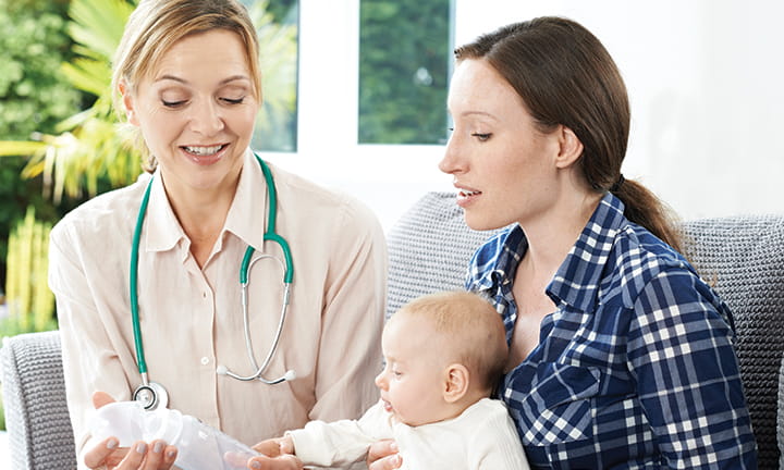 mom holding newborn sitting on couch with doctor who is showing her a baby bottle