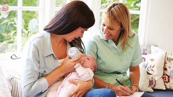 mom feeding infant a bottle while nurse watches and takes notes