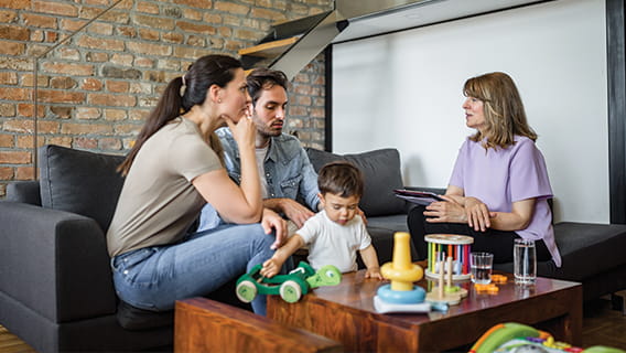 mom and dad sitting on couch talking to nurse as infant plays with toys next to them