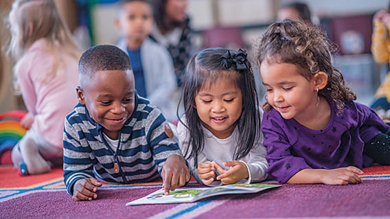 three preschoolers laying on floor in class looking at a book together