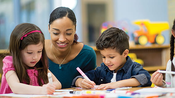 preschool teacher kneeling and smiling at a table with two students while they write