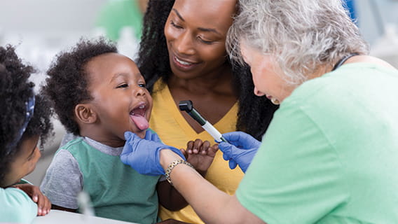 mom holding toddler son while nurse checks his tongue and toddler sister watches