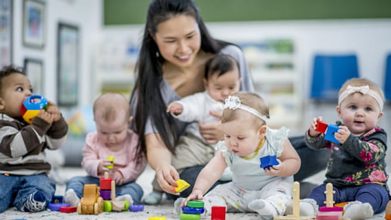 provider sitting on floor in care setting holding a baby  surrounded with two other babies playing with toys
